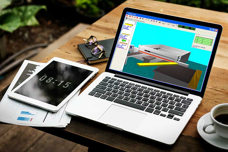 Egy fa asztalon kávé, szemüveg, jegyzetfüzet, kinyomtatott diagrammokon egy tablet és egy laptop. A laptopon az Ottimo szoftverben egy 3D szimuláció fut.