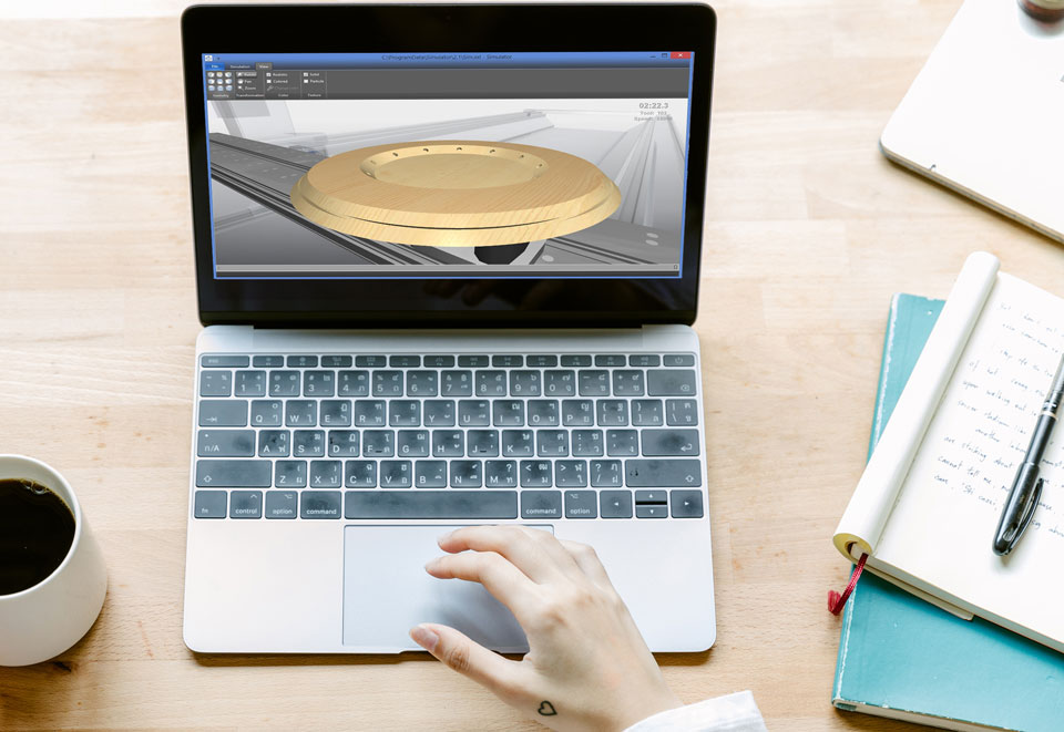 Egy fa asztalon jegyzetfüzetek, kávé és középpen egy laptop, amin egy hölgy kéeszül gépelni, kezén egy szivecske tetoválással. A laptopon egy munkadarab 3D-s előnézete látható.