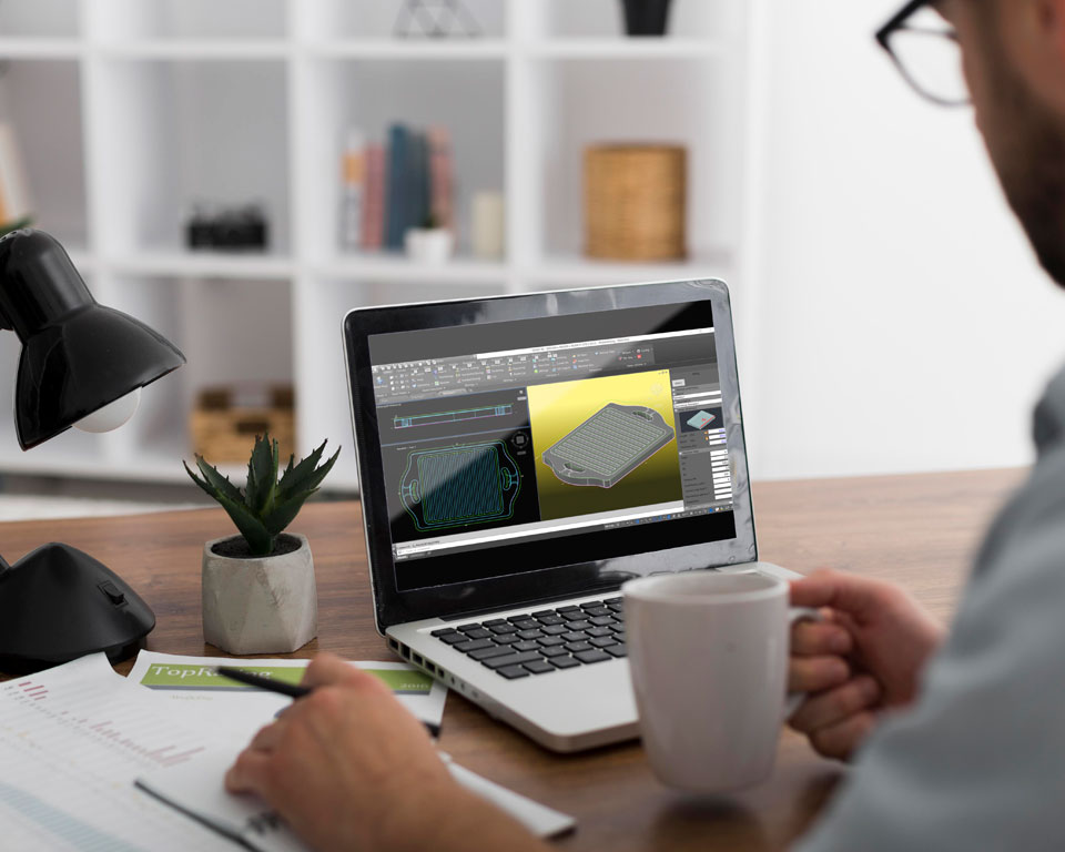 Egy fehér irodában egy férfi ül az íróasztal mögött egyik kezében egy tollal, a másikban egy bögrével, és az előtte, egy fa asztalon lévő lapton egy 3D CAD rajzot tanulmányoz.