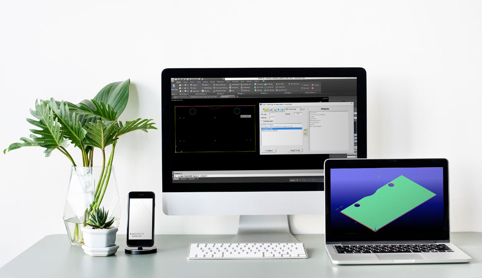 Egy fehér fal előtt lévő asztalon egy mobiltelefon, egy iMac és egy laptop látható. Mindhárom kijelzőjén CAD-es felülettel.